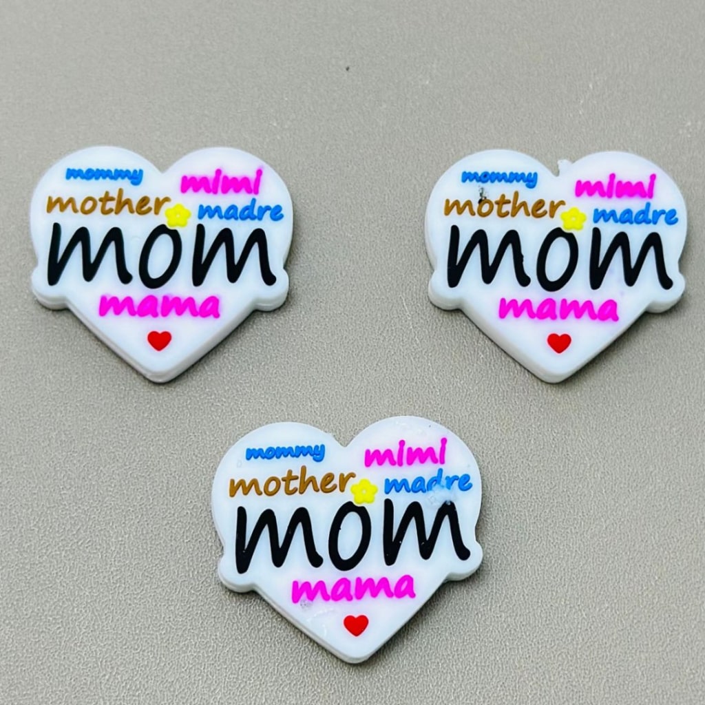 Heart-shaped silicone beads with the word 'mom' and variations like 'mother', 'mama', and 'mummy' written on them in different colors.

