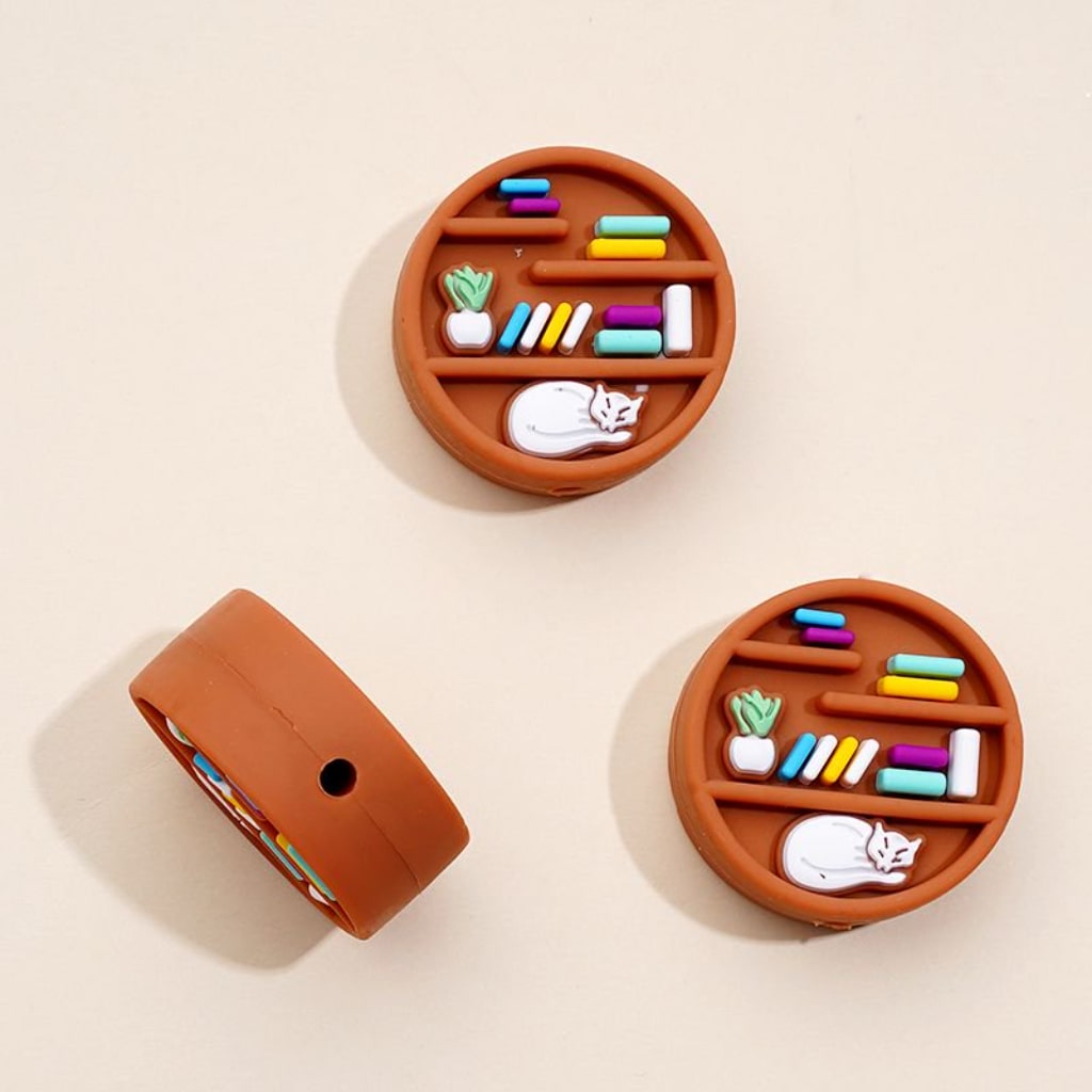 Three brown silicone beads designed to look like bookshelves with colorful books on them, displayed against a beige background.

