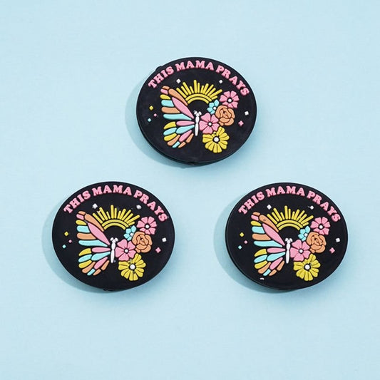 Three round silicone beads with a colorful butterfly and flower design and the text 'MAMA PRAYS' on a black background.

