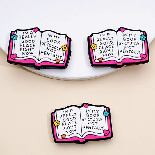 Silicone beads in the shape of an open book with the phrase 'In a Really Good Place Right Now In My Book Of Course Not Mentally' printed on them, in pink, white, black, and multicolor.

