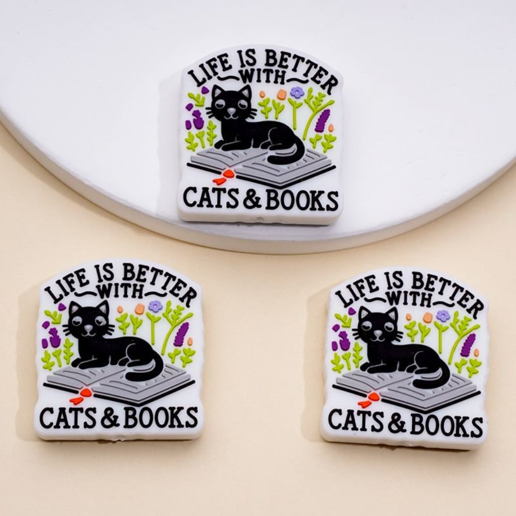 Silicone beads featuring a black cat sitting on a book with the phrase 'LIFE IS BETTER WITH CATS & BOOKS' printed above.

