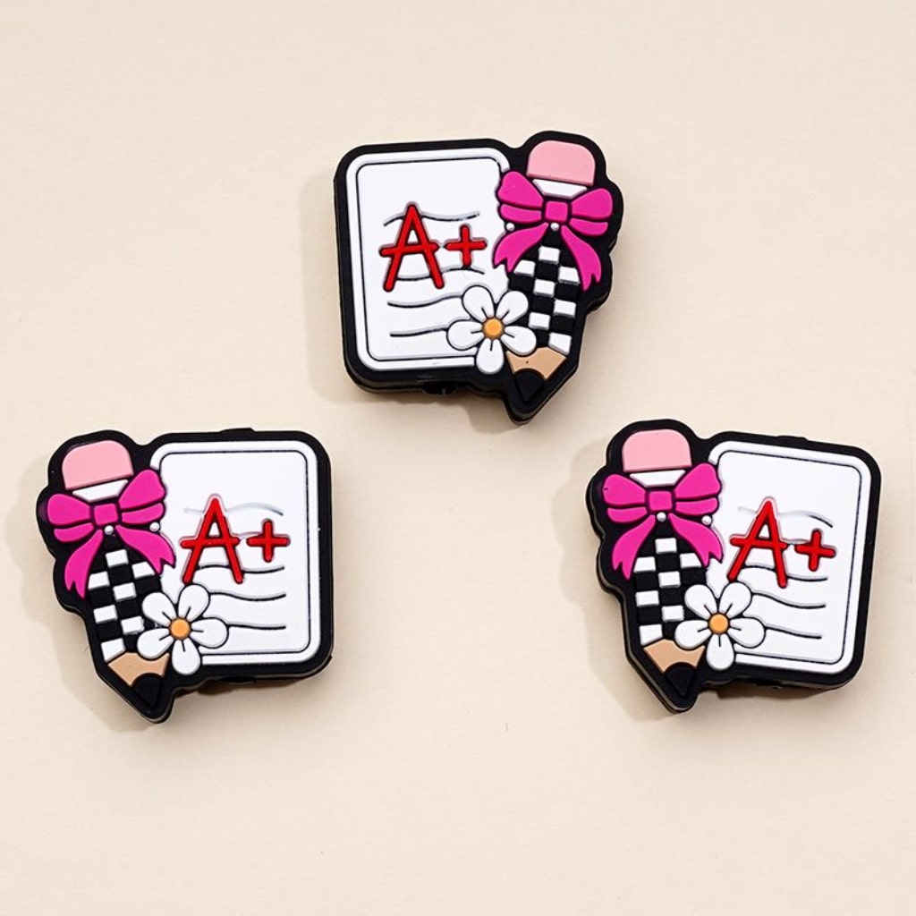 Three silicone beads designed as pencils with flowers and a plus sign, against a white background.

