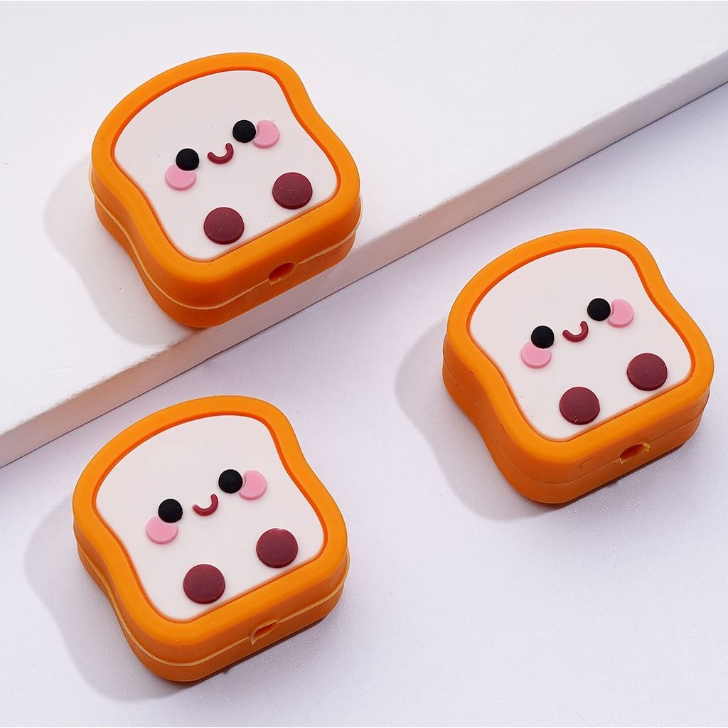 Silicone beads shaped like pieces of toast with smiling faces and eyes, in orange, white, and black colors.

