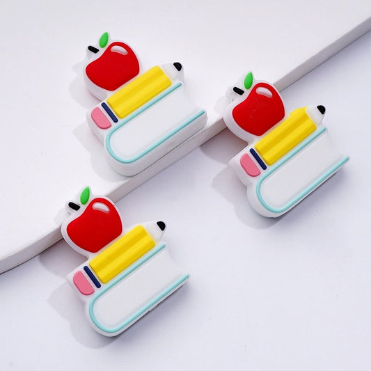 The image shows three silicone beads in the shape of a red apple, a yellow pencil, and a white book, each with a simple and playful design.

