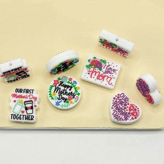 A collection of silicone focal beads with various designs including floral patterns and text that says 'Happy Mother's Day' and 'I love you mom'. The beads are primarily white and red with colorful accents.


