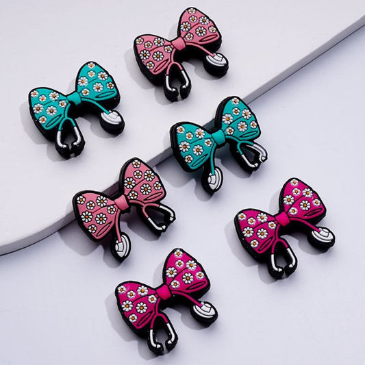 A collection of silicone beads in the shape of bowknots with mini flowers, in black, pink, and teal colors, with metallic clasps for attaching to stethoscopes or crafts.

