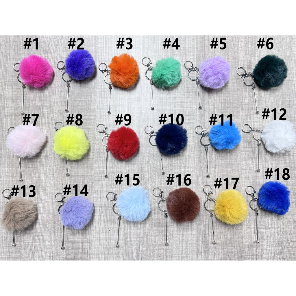 A collection of fluffy pom pom balls with metal keychains in various colors attached to a beige surface.

