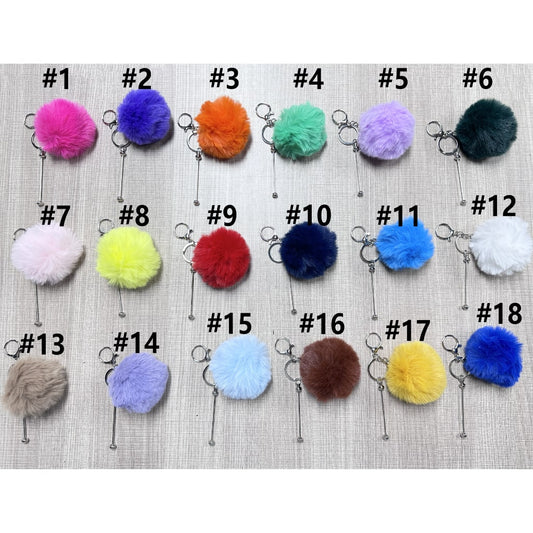 A collection of fluffy pom pom balls with metal keychains in various colors attached to a beige surface.

