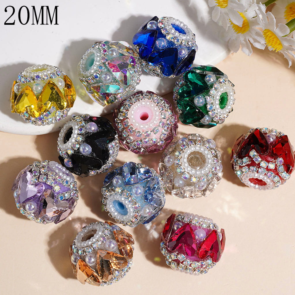 Ten 20mm faceted crystal beads in various colors and rhinestone patterns, arranged on a light beige surface with white daisies in the corner.

