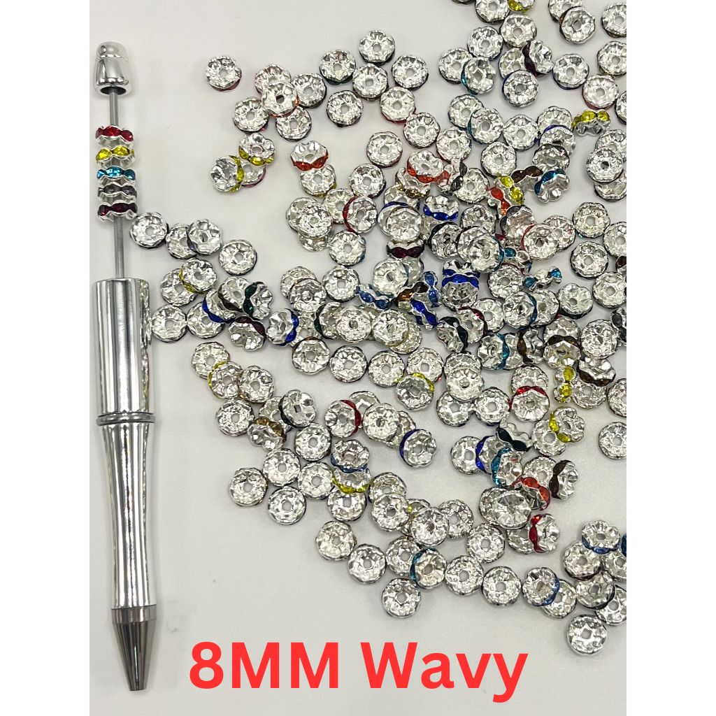 A collection of round spacers with wavy shapes and rhinestones, in various colors, displayed on a surface with a pen for scale.

