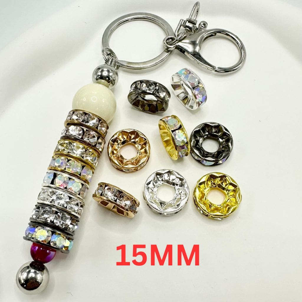 A collection of round metal spacers with large holes, some adorned with rhinestones, displayed on a white surface. The size of the spacers is marked as 15mm.

