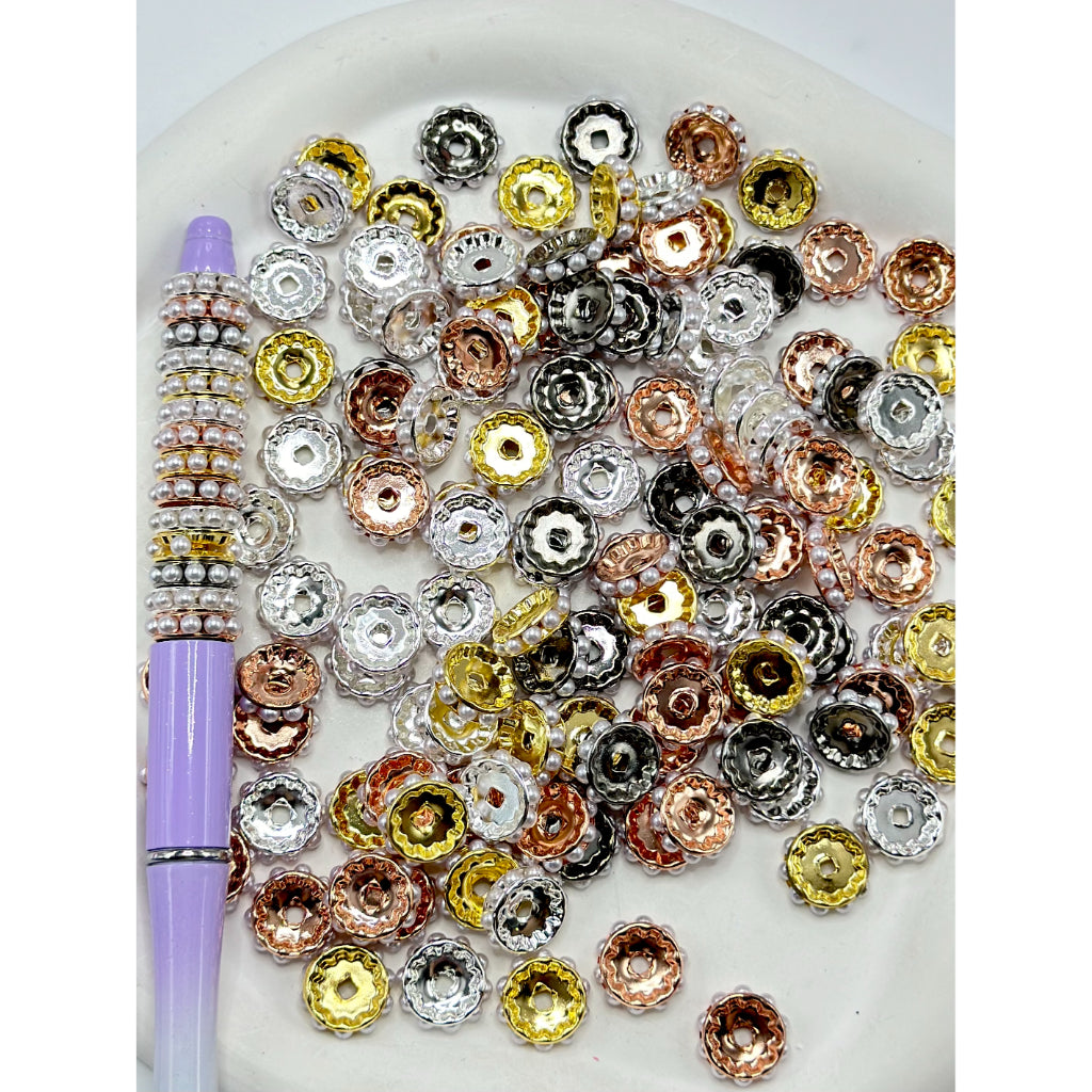 A collection of metal beadable spacers with white pearls in a variety of colors including silver, gold, and rose gold, displayed on a plate with a pen for scale.

