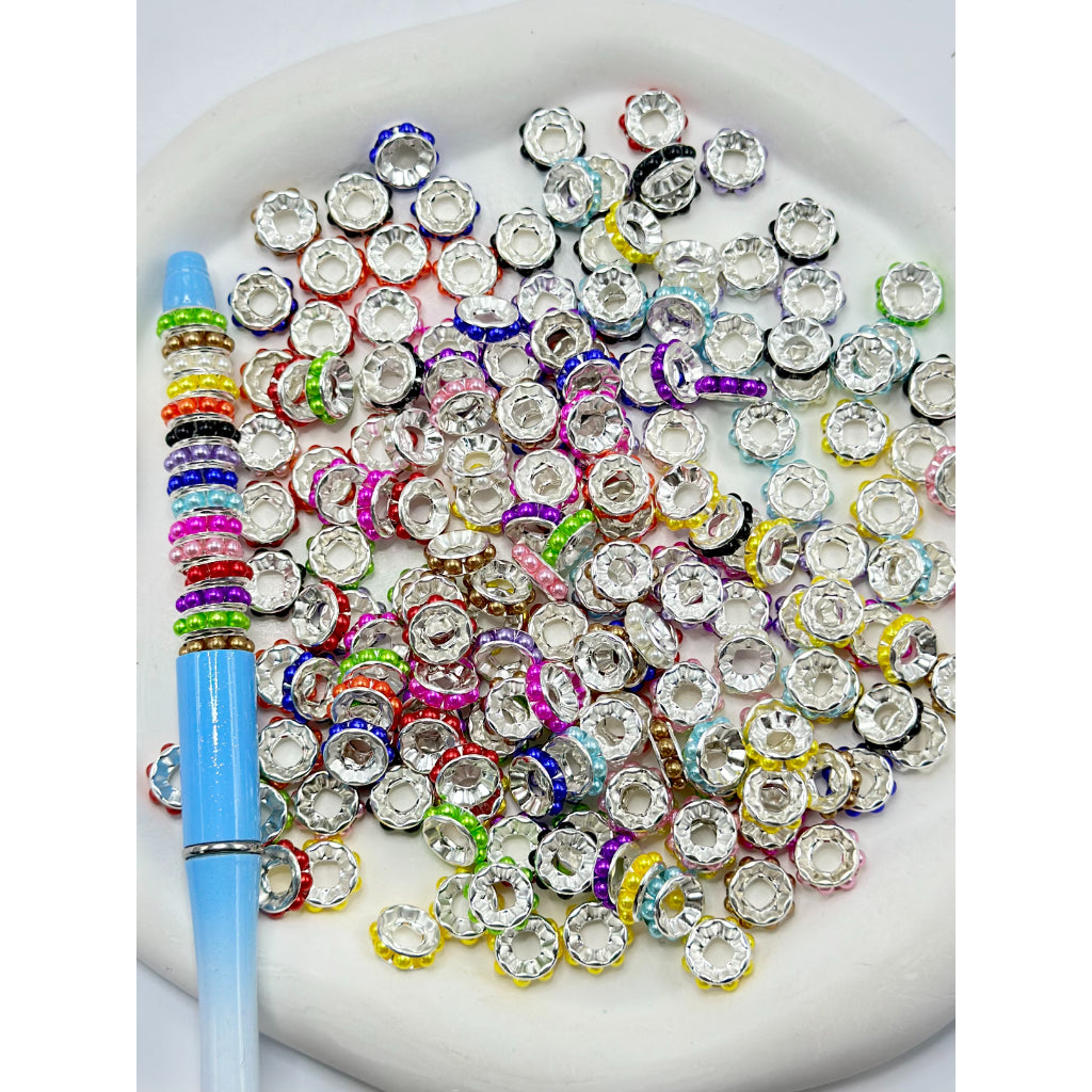 A collection of multicolored beadable metal spacers with pearls, arranged on a surface with a pen for scale.

