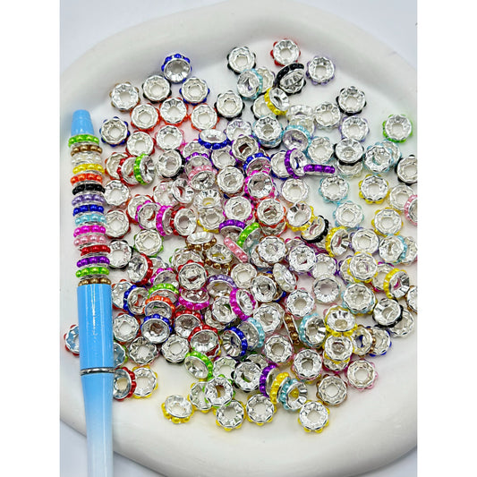 A collection of multicolored beadable metal spacers with pearls, arranged on a surface with a pen for scale.

