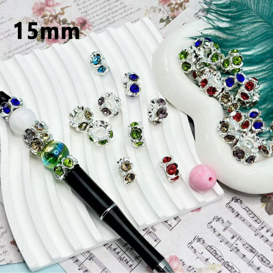 Delicate silver metal spacers with flower designs and colorful rhinestones inside, displayed on a white surface next to a pen for scale.

