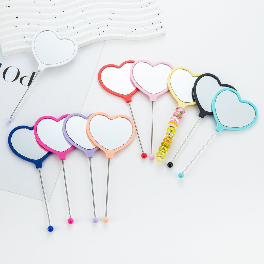 A collection of heart-shaped handheld makeup mirrors in various solid colors, each with a different color bead attached to the end.

