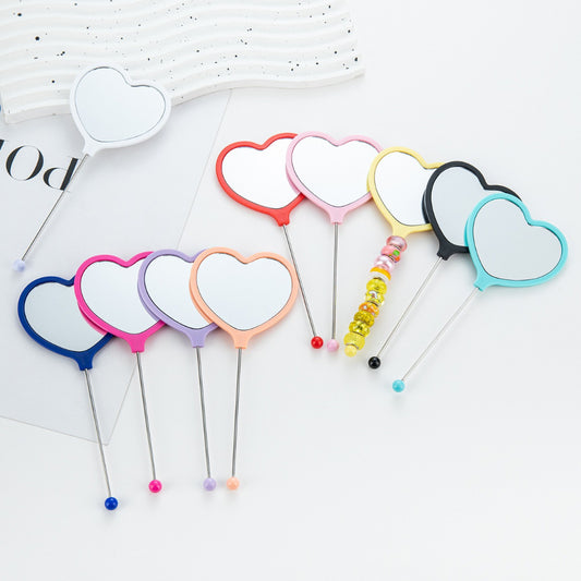A collection of heart-shaped handheld makeup mirrors in various solid colors, each with a different color bead attached to the end.

